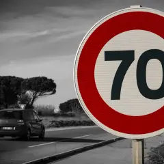 Zbliża się znak ograniczenia prędkości do 70 km/h. Samochód jedzie po drodze w tle.