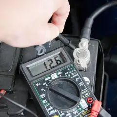 Dłoń z multimetrem sprawdza napięcie akumulatora. Odczyt 12.6V pokazuje, jak sprawdzić akumulator.