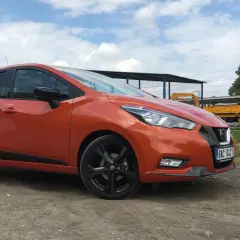Pomarańczowy Nissan Micra na tle torów kolejowych. Sprawdź nissan micra wymiary, by poznać jego kompaktowe gabaryty.
