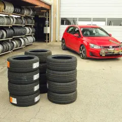 Czerwony Volkswagen Golf GTI z zamontowanym sprzętem do testu opon letnich, obok stosy nowych opon i regały z felgami.