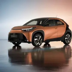 Pomarańczowy Aygo X Hybrid z czarnymi akcentami i stylowymi felgami, odbijający się w lśniącej podłodze.