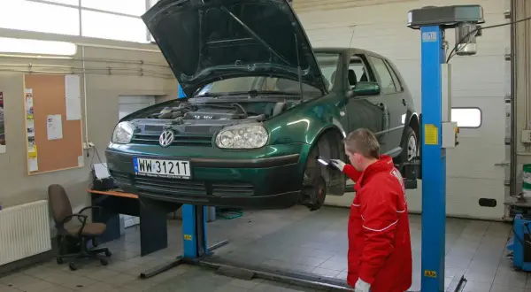 Mechanik w czerwonej kurtce sprawdza podniesiony na podnośniku zielony samochód. To kluczowy moment, by dowiedzieć się, jak dbać o samochód.