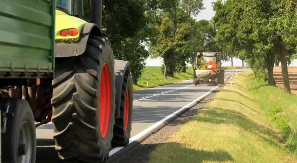 Ciągnik z przyczepą przygotowuje się do wyprzedzania pojazdu wolnobieżnego na wiejskiej drodze.