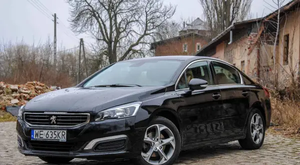 Czarny Peugeot 508 2.0 HDI na brukowanej drodze, w tle zniszczone budynki i drzewa.