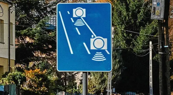 Niebieski znak drogowy z białymi symbolami aparatów fotograficznych i falami, informujący o odcinkowym pomiarze prędkości.