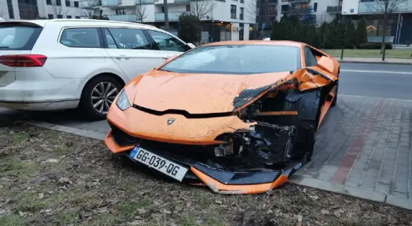 Pomarańczowe lambo po wypadku, przód mocno zniszczony. Obok białe kombi.
