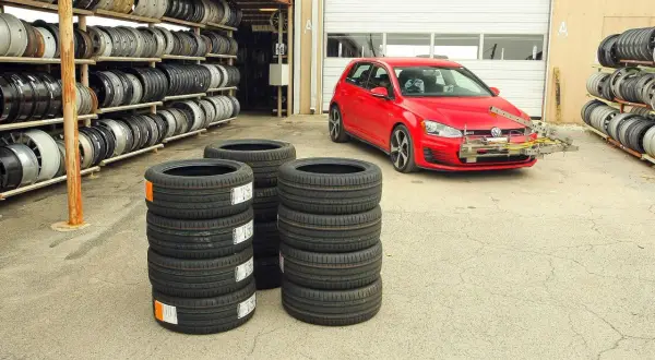 Czerwony Volkswagen Golf GTI z zamontowanym sprzętem do testu opon letnich, obok stosy nowych opon i regały z felgami.