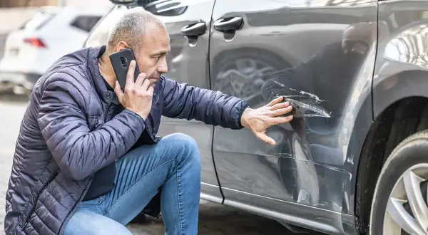 Mężczyzna sprawdza uszkodzenia na samochodzie po stłuczce samochodowej nie z jego winy. Dzwoni po pomoc, wskazując na głębokie rysy.