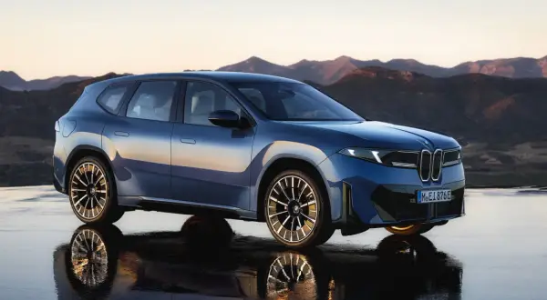 Niebieski BMW Neue Klasse, SUV z futurystycznym designem, stoi na mokrej nawierzchni z odbiciem gór w tle.