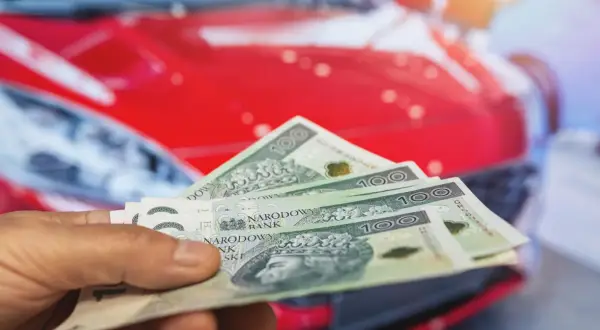Dłoń trzyma banknoty 100 zł na tle czerwonego samochodu. Koszty posiadania auta, w tym podatek od samochodu, mogą być znaczące.