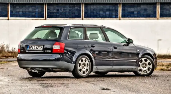 Ciemnogranatowe Audi A4 B6 1.9 TDI Avant stoi na mokrym asfalcie przed budynkiem z niebieskimi oknami.