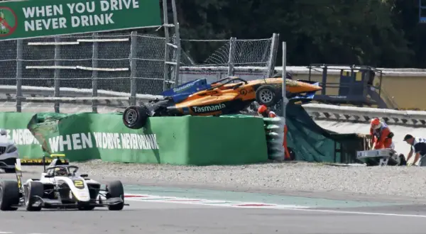 Wypadek bolidu F1 na torze Monza. Pomarańczowo-niebieski bolid leży na bandzie, ratownicy udzielają pomocy.