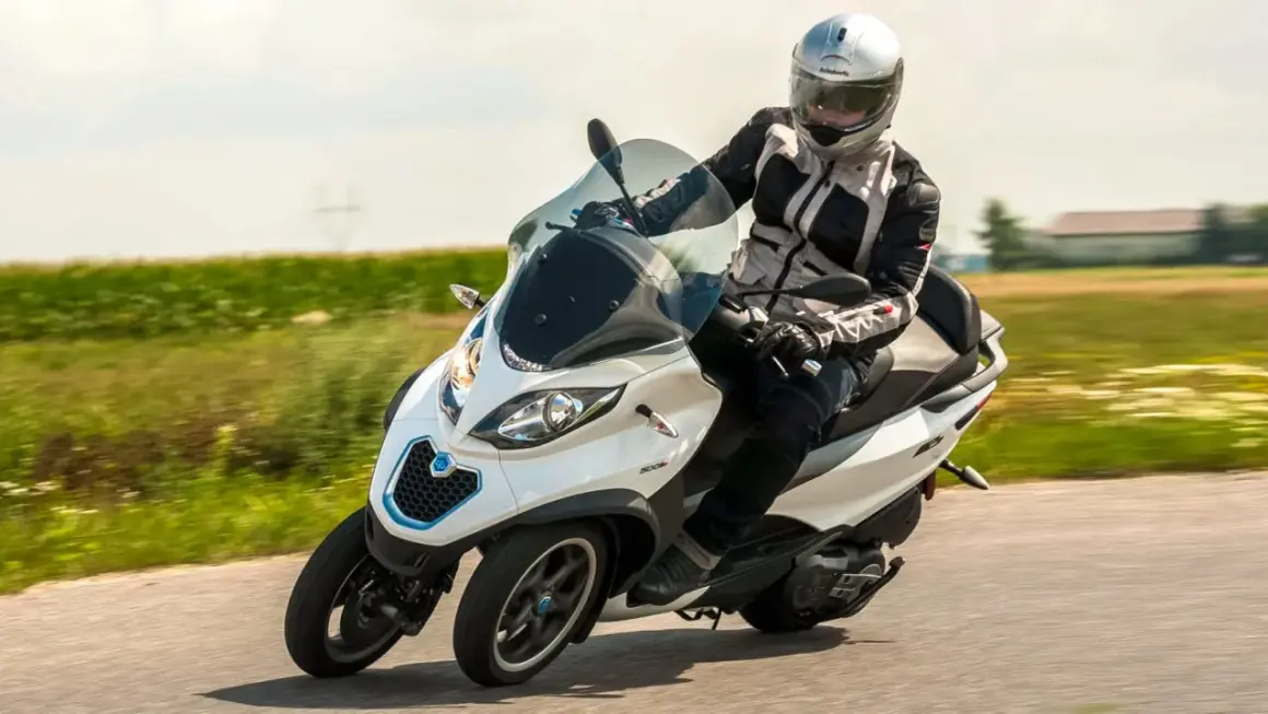 Kierowca w kasku na białym Piaggio MP3 500, pokonujący zakręt na drodze. Czy wady tego modelu są widoczne?