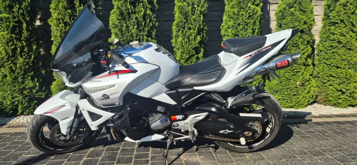 Biały Suzuki B-King z czarnym siedzeniem i sportowym wydechem Yoshimura, zaparkowany na kostce brukowej, na tle zielonych żywopłotów.