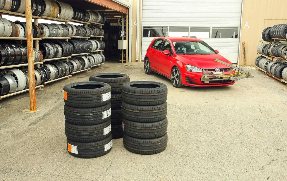 Czerwony Volkswagen Golf GTI z zamontowanym sprzętem do testu opon letnich, obok stosy nowych opon i regały z felgami.