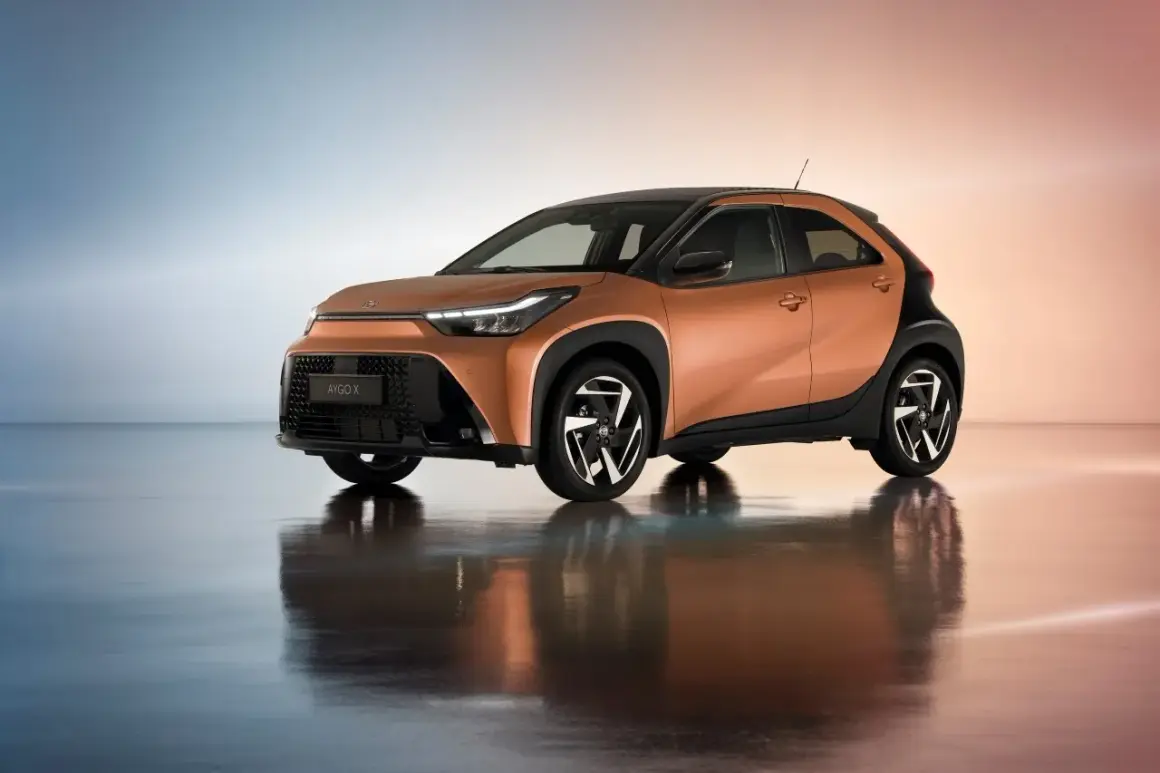 Pomarańczowy Aygo X Hybrid z czarnymi akcentami i stylowymi felgami, odbijający się w lśniącej podłodze.