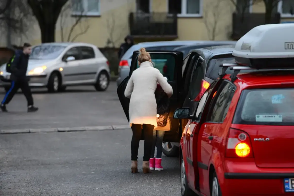 Kobieta z dzieckiem wsiada do samochodu, tworząc zator. Miejsce, gdzie nie można parkować, bo blokuje ruch.