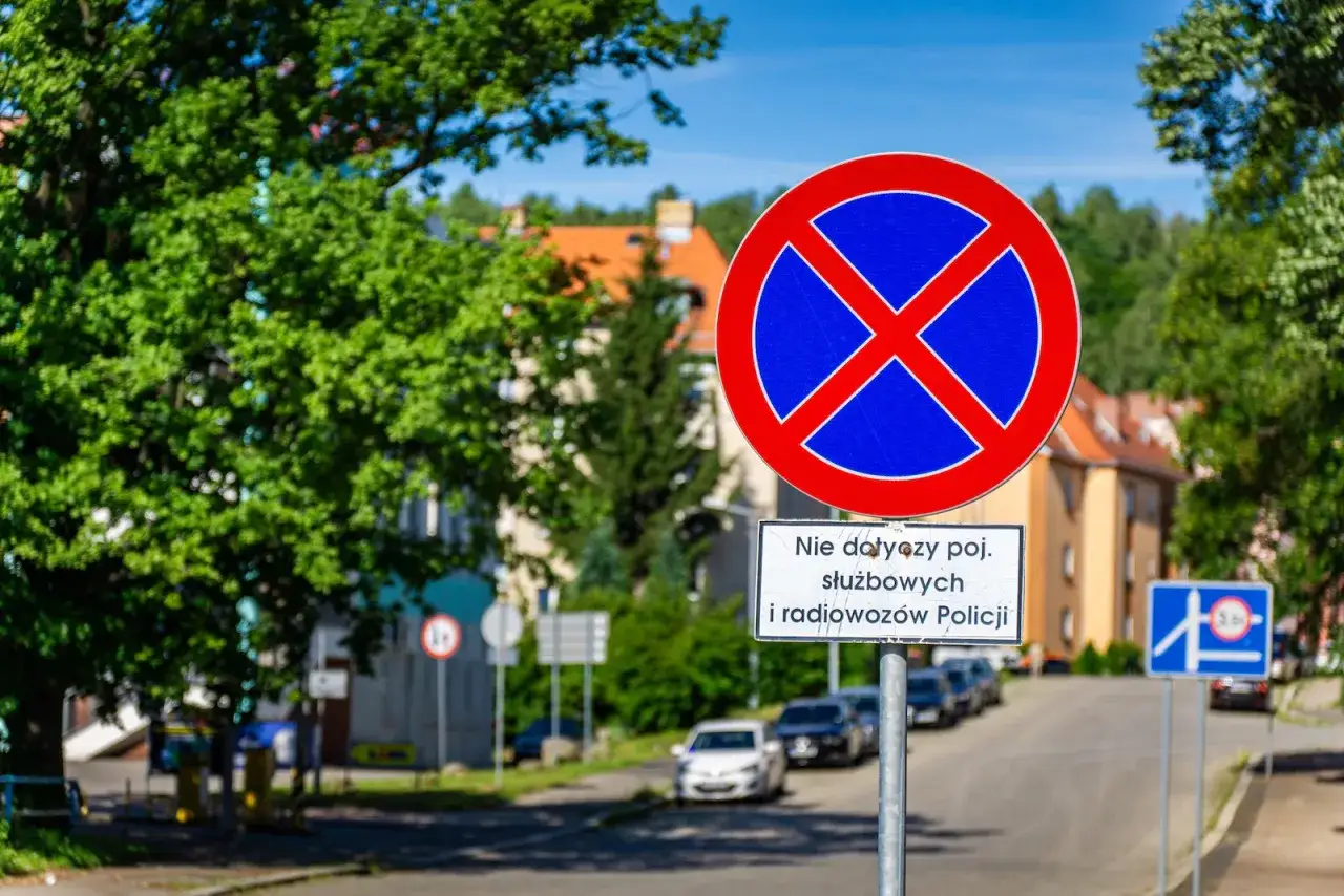 Znak zakazu zatrzymywania się informuje, gdzie nie można parkować. Dodatkowa tabliczka precyzuje, że zakaz nie dotyczy pojazdów służbowych i radiowozów Policji.