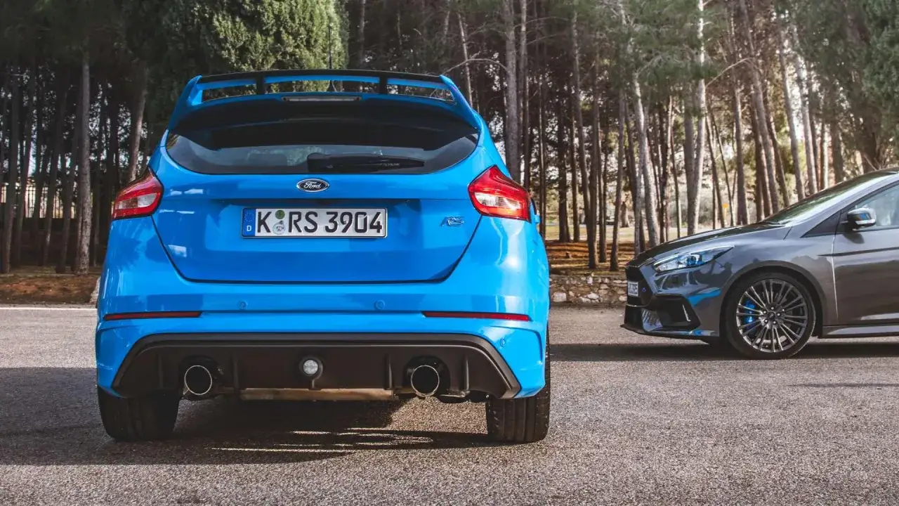 Dwa Fordy Focus RS: jeden niebieski z tyłu, drugi szary z przodu, zaparkowane na tle lasu.