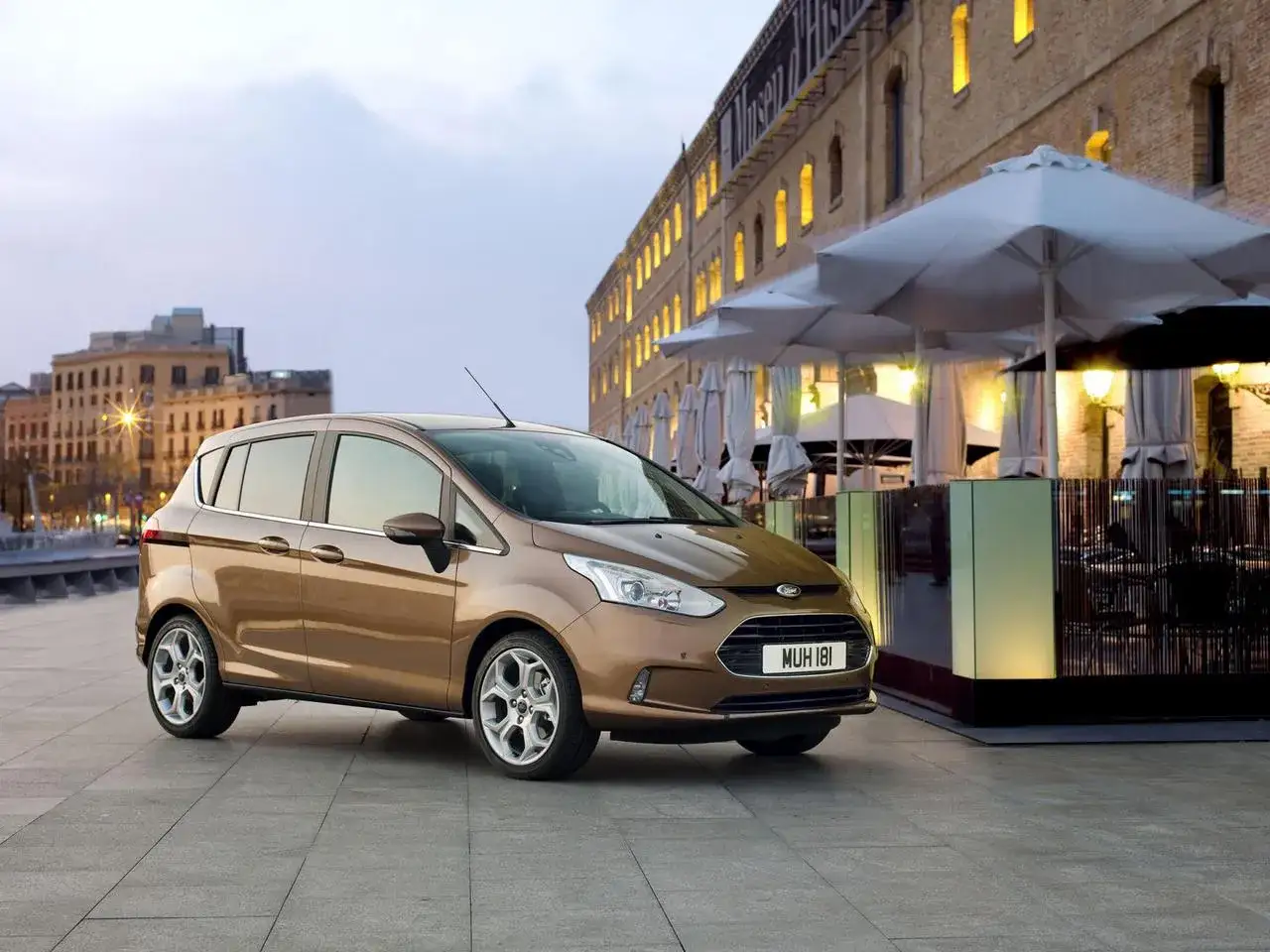 Brązowy Ford B-Max na tle miejskiej zabudowy. Zastanawiasz się nad jego wymiarami? Ten kompaktowy samochód oferuje zaskakująco dużo przestrzeni.