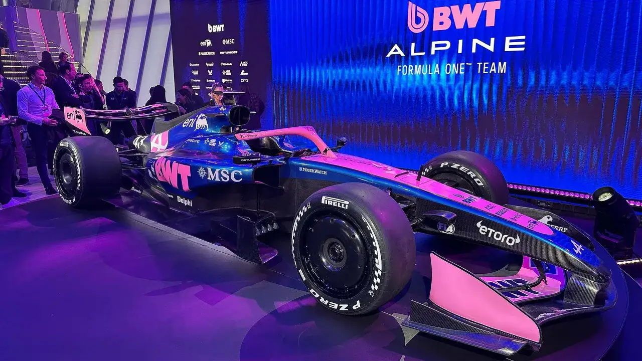 BWT Alpine F1 Team bolid w kolorach niebieskim i różowym, prezentowany na tle niebieskiego ekranu z logo zespołu.