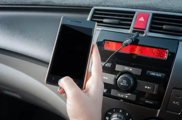 Dłoń trzyma smartfon podłączony kablem do radia samochodowego. Wybrano wejście aux.
