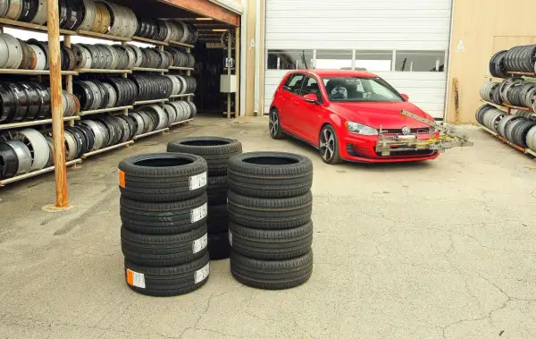 Czerwony Volkswagen Golf GTI z zamontowanym sprzętem do testu opon letnich, obok stosy nowych opon i regały z felgami.