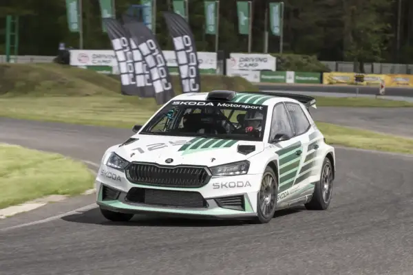 Biała Skoda Fabia Rally z zielonymi pasami pędzi po torze. Kierowca w kasku jest gotowy do akcji.