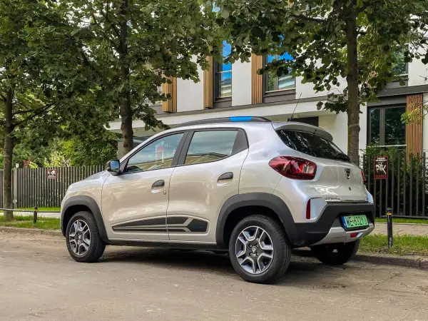 Srebrna Dacia Spring zaparkowana pod drzewami. Ten elektryczny samochód oferuje dobry zasięg na miejskie podróże.